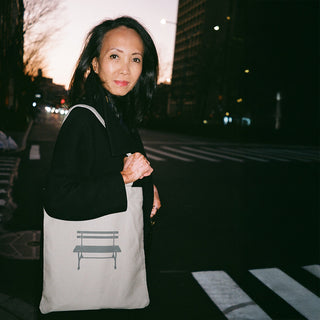 a person crosses a NYC street at dusk while carrying a Light grey tote with a grey bench printed on it