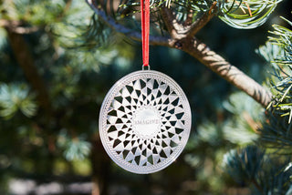 a silver-colored metal ornament in the design of the Central Park Imagine Mosiac with a red satin ribbon hanging on an evergreen tree