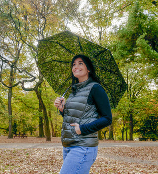 Woman smiling under a green-patterned canopy umbrella in Central Park surrounded by autumn trees. All products available through Central Park Conservancy Shop.