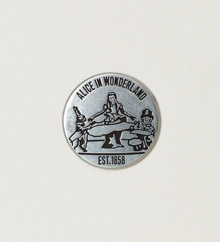 Close-up of a round silver token featuring Alice in Wonderland characters with 'Alice in Wonderland, Est. 1858' text on a white background.