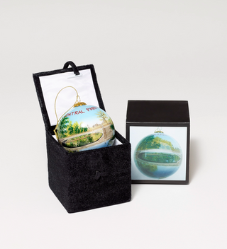 Central Park-themed ornament with a bridge and pond scene, partially unpacked from a black velvet box.