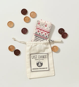 a folded printed cream, red and grey bandana emerging from a printed muslin drawstring bag and scattered light and dark wooden checkers on a  grey background