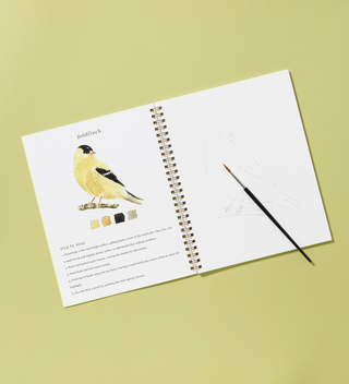 Birds watercolor workbook, open to the goldfinch page with a watercolor brush on top