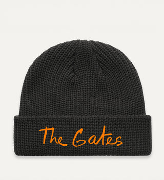 Black beanie with The Gates in Christo's handwriting embroidered in saffron orange thread on a grey background.