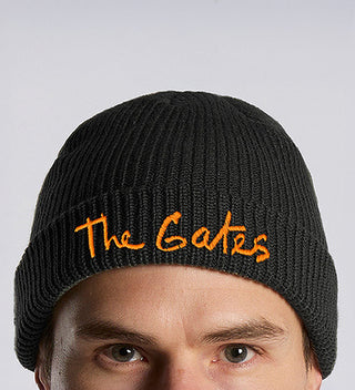 a closeup of a model  wearing a black knit beanie with The Gates in Christo's signature handwriting embroidered in saffron orange thread. 