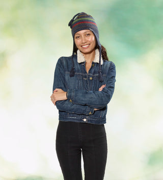 a  smiling woman with her arms crossed wears a striped navy knit hat with earflaps, top tassel and tassel ties and a woven wrap-around label with 'Central Park Est. 1858, NYC' in front of a green dappled background