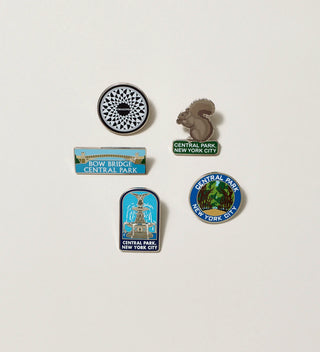 Close up of five enamel pins on a white background, including two round pins, a horizontal rectangular  'Bow Bridge' pin, a vertical rectangular 'Fountain' pin and a squirrel-shaped pin.