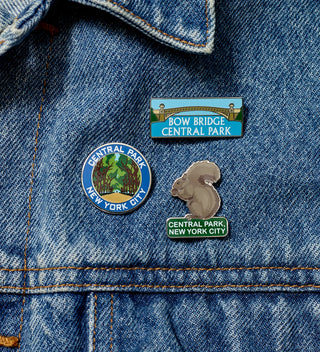 Three Central Park-themed pins attached to a denim fabric, including a round pin, a rectangular 'Bow Bridge' pin, and a squirrel-shaped pin.