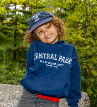 a smiling child in Central park wears an 1858 hoodie while sitting on a boulder