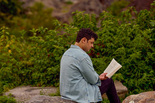 Man wearing a blue denim shirt is sitting on a rock in Central Park and reading a book