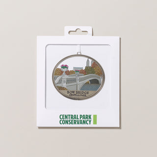 Bow Bridge Central Park ornament with a detailed design of the bridge and surrounding foliage in a flat white envelope with a window and the Central Park Conservancy logo on a gray background.