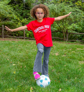 a childing playing with a soccer ball in Central Park  wears a red t-shirt with Central Park , New York City, Since 1858 design in white ink 