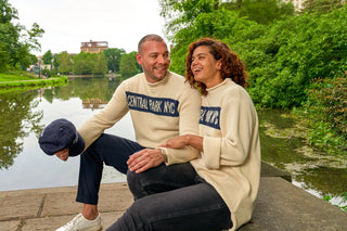 two  smiling people sit on in Central  Park wearing cream colored roll neck sweaters with CENTRAL PARK  NYC in Blue banner across the chest. 