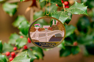 A closeup of an oval laser cut metal ornament of Bow Bridge in Central Park hanging in a holly tree 