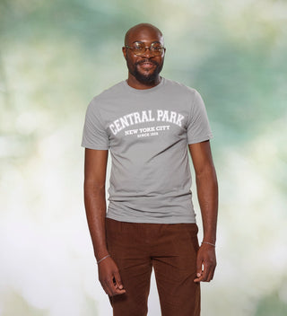 Man standing and smiling, wearing a gray t-shirt with 'Central Park New York City Since 1858' text, against a muted nature  background.