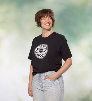 Woman wearing a black t-shirt with a circular 'Imagine' mosaic design, inspired by John Lennon, smiling and posing with her hand in her pocket.