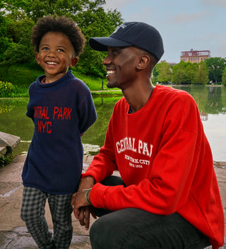 a smiling child  wears a navy sweater with red text reading Central Park NYC next to a squatting man with a red sweatshirt and blue hat