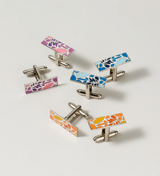 Assorted Central Park Conservancy cufflinks in different colors and designs, with a packaged pair next to a black stone on a light grey surface