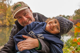 a father wearing a tan Conservancy logo hat and a child wearing a blue melange knit hat embracing in Central Park