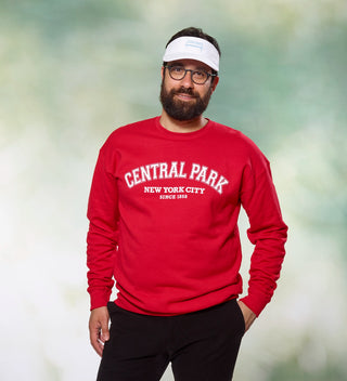 Man posing while wearing a red "Central Park New York City" sweatshirt and white visor with a Central Park bench