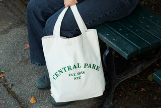A closeup  shot of seated person holding a natural canvas tote bag with  Central Park Est.1858 NYC printed on it in dark green 