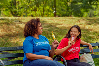 Image of two women laughing on Central Park bench wearing t-shirts sold at the Central Park Conservancy shop