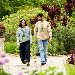 two people walking in Central Park Conservancy Garden, one wearing a stone colored crewneck sweatshirt featuring a green screenprint of Bethesda Fountain and the Central Park Conservancy word mark.  