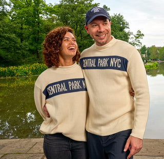 Two  smiling people sit on in Central  Park wearing cream colored roll neck sweaters with CENTRAL PARK  NYC in Blue banner across the chest. 