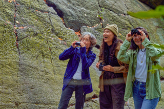 three people use binoculars with a large boulder as a background