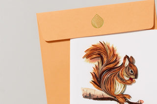 A close-up of a square card featuring a quilled paper design of a squirrel in browns and oranges and a golden yellow envelope 