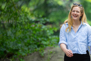 a smiling woman in a blue linens shirt, dangling blue Minton earrings  and sunglasses on her head walks in Central Park