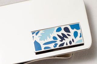 a close-up photograph of a silver money clip with a blue leaf and botanical design