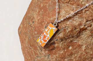 a closeup of a necklace with an orange leaf and botanical design laying on a natural orange stone