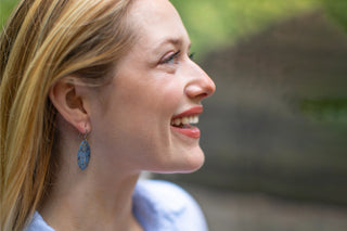 a  smiling woman wears blue and silver dangle earrings  with ornate designs inspired by the Minton tiles in Bethesda Terrace