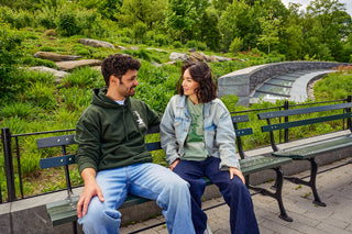 A man and a woman sitting on a green Central Park bench, smiling at each other. The background features the lush green roof at the Davis Center in Central Park, NYC.