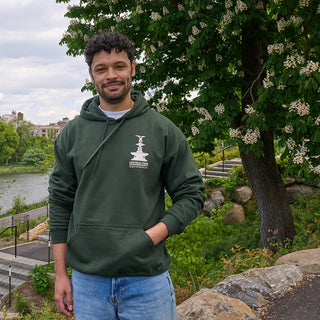 Man wearing a forest green hoodie with a logo standing near the Davis Center in Central Park among trees.