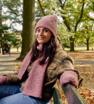 Woman in a cozy red knit hat and scarf sitting on a park bench in Central Park surrounded by autumn leaves. All products available through Central Park Conservancy Shop.