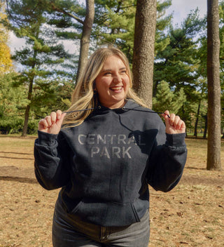 Woman wearing a monochrome black 'Central Park' hoodie, smiling in Central Park with tall trees in the background. All products available through Central Park Conservancy Shop.