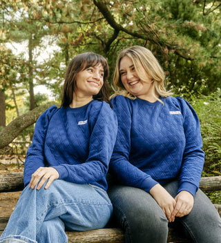 Two women in matching blue quilted puff-sleeve pullovers sitting on a wooden bench in Central Park. All products available through Central Park Conservancy Shop.