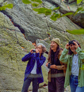 Three adults enjoying birdwatching in a scenic, rocky area in The Ramble. Two people hold binoculars, and all are smiling, capturing the essence of outdoor exploration and camaraderie.