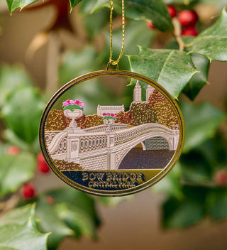 Circular brass ornament depicting Bow Bridge in Central Park, surrounded by lush greenery and intricate details. All products available through Central Park Conservancy Shop.