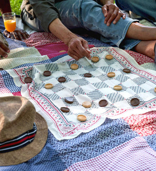 Close-up of a group playing checkers on a patchwork quilt during a picnic in the park. The checkers board is laid on a patterned cloth, with hands moving pieces.