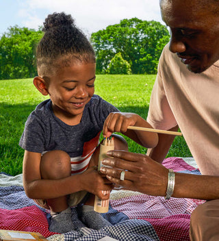 Young child sitting on a picnic blanket, smiling as they hold colored pencils, assisted by an adult, while enjoying a sunny day in Central Park.