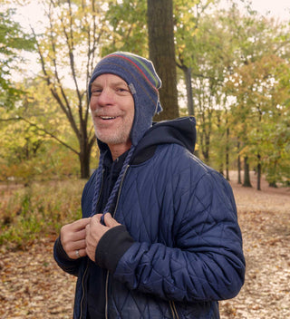 Man wearing a colorful knit ear-flap hat, smiling in Central Park's wooded area. All products available through Central Park Conservancy Shop.