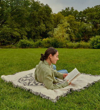 A young woman lying on a blanket with a design resembling the 'Imagine' mosaic, reading a book while enjoying a quiet moment in Central Park’s grassy area.