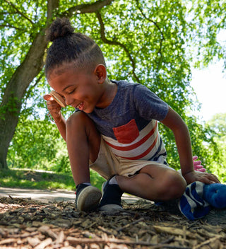 Young child laughing, crouched down with a toy dual-magnifier in hand, surrounded by nature under a large tree in Central Park.