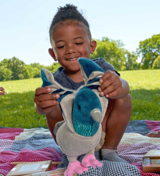 Child smiling while holding a soft, plush pigeon toy during a picnic in Central Park, sitting on a colorful patchwork blanket.