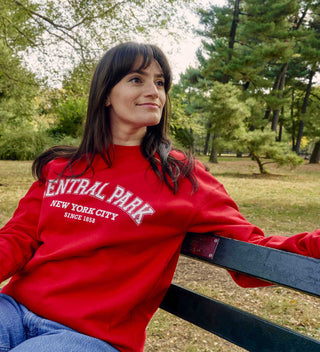 Woman wearing a bright red 'Central Park, New York City, Since 1858' sweatshirt, sitting on a park bench in Central Park surrounded by trees. All products available through Central Park Conservancy Shop.