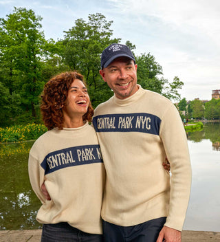 Couple smiling and standing by the Harlem Meer in Central Park, both wearing matching 'Central Park NYC' rollneck sweaters.