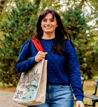 Woman walking through Central Park carrying a canvas tote bag illustrated with a vibrant map of the park and landmarks. All products available through Central Park Conservancy Shop.